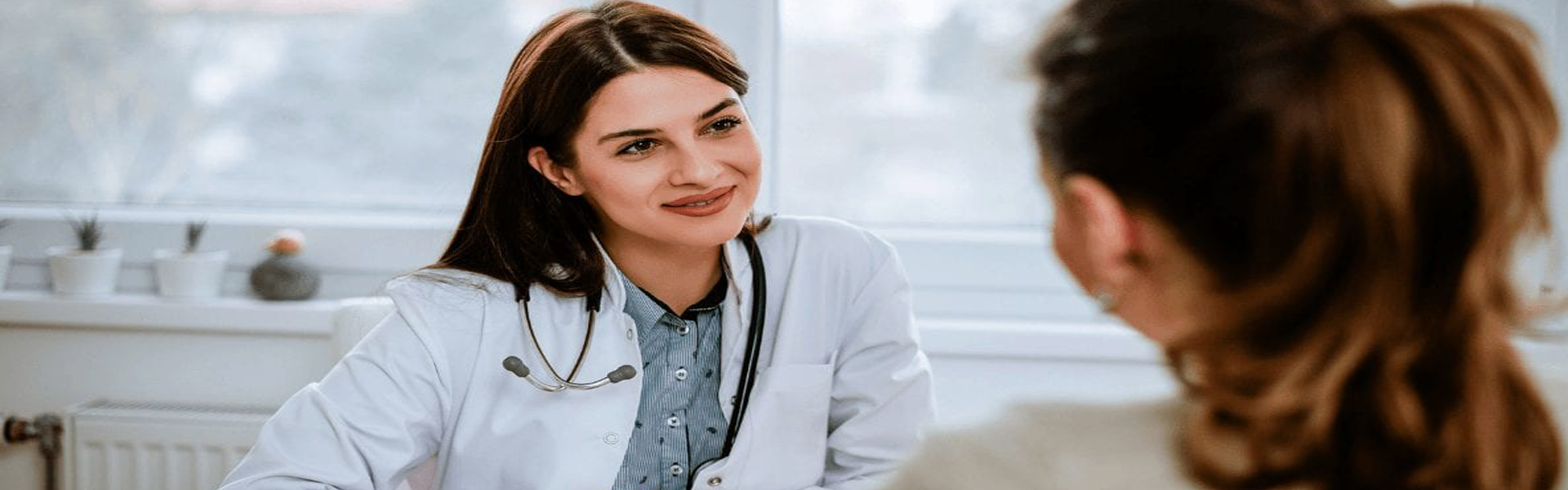 Picture of a female doctor consulting with a patient.  The doctor is wearing a white smock, sitting at a desk, and talking with a patient sitting in front of her.